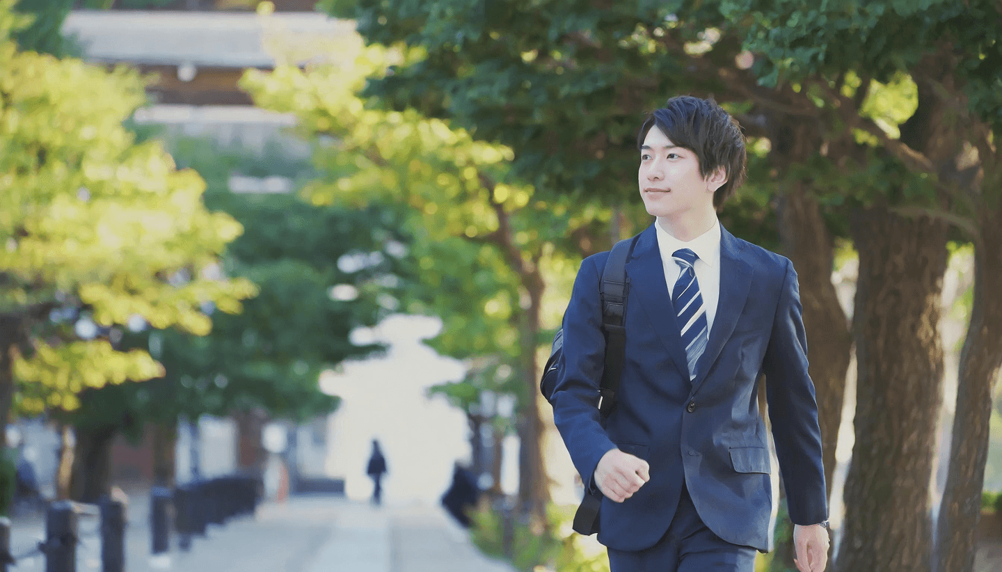 スーツ姿で歩く若い男性、緑に囲まれた通りを笑顔で前向きに進む様子